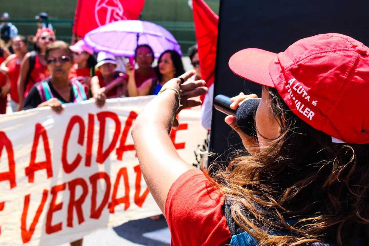 Manifestação pelo direito à moradia: pessoa fala ao microfone defronte a manifestantes exibindo faixa