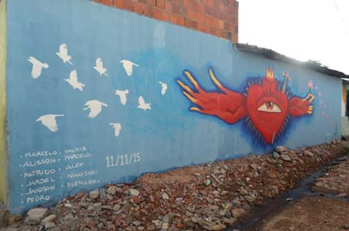 Ilustração sobre muro azul mostra um coração vermelho à direita, com um olho ao meio e de onde saem dois braços, com 11 pássaros brancos à esquerda e os nomes das vítimas no canto inferior esquerdo 
