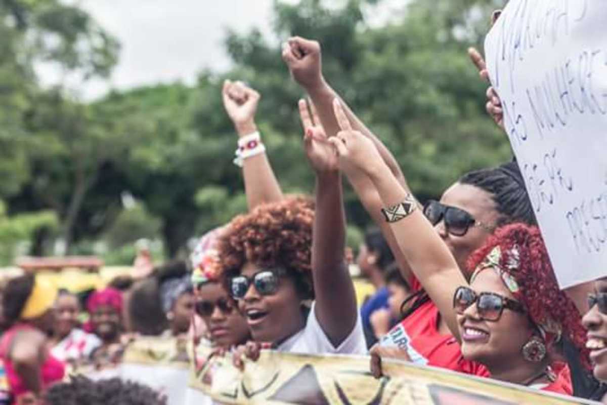 Mulheres e homens negros de mãos erguidas ou com faixas e cartazes durante manifestação pública