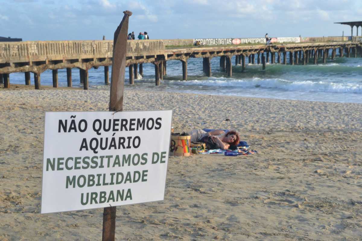 Manifestante contra Aquario Ceará, deitado na areia da Praia de Iracema, com o mar e um espigão ao fundo e em primeiro plano uma placa com os dizeres 