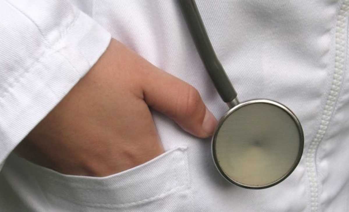 Imagem em detalhe de um profissional de saúde,  com mão no bolso e parte do estetoscópio junto a ela
