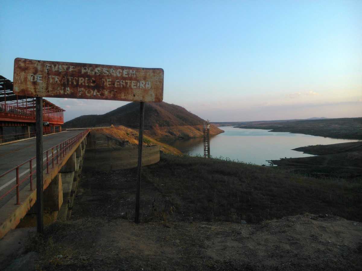 Uma placa de sinalização deteriorada, uma ponte e um açude com pequeno volume de água: cena do Interior do Ceará