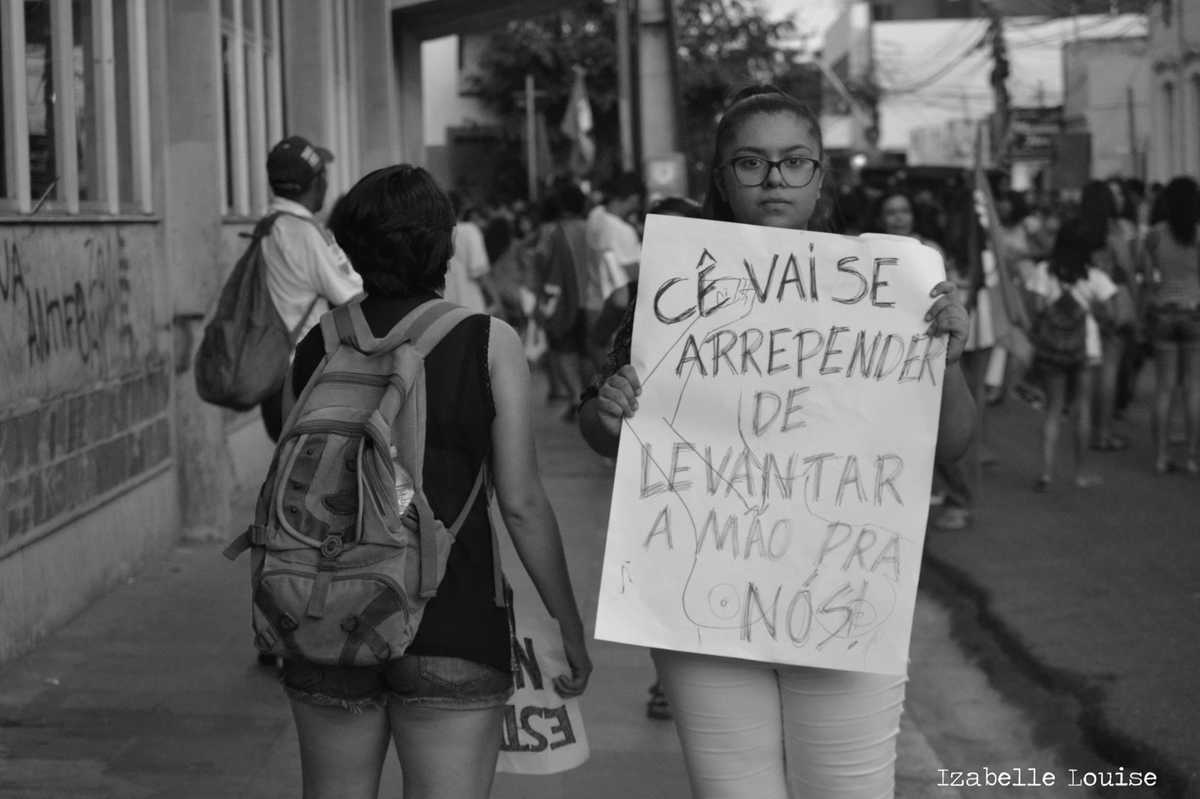 Mulher jovem, na rua, com pessoas circulando ao fundo, segura um cartaz com os dizeres 