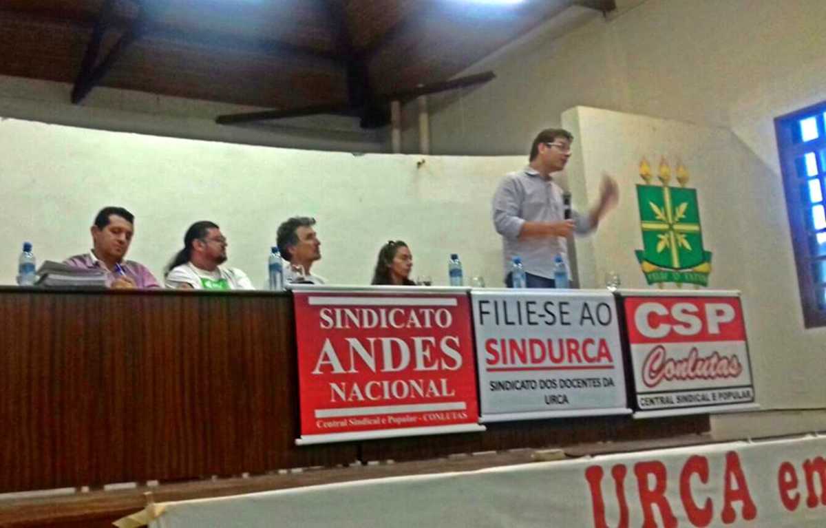 Renato Roseno, em pé, fala em mesa com outras quatro pessoas sentadas e cartazes do Sindicato Andes Nacional, Sindurca e CSP Conlutas