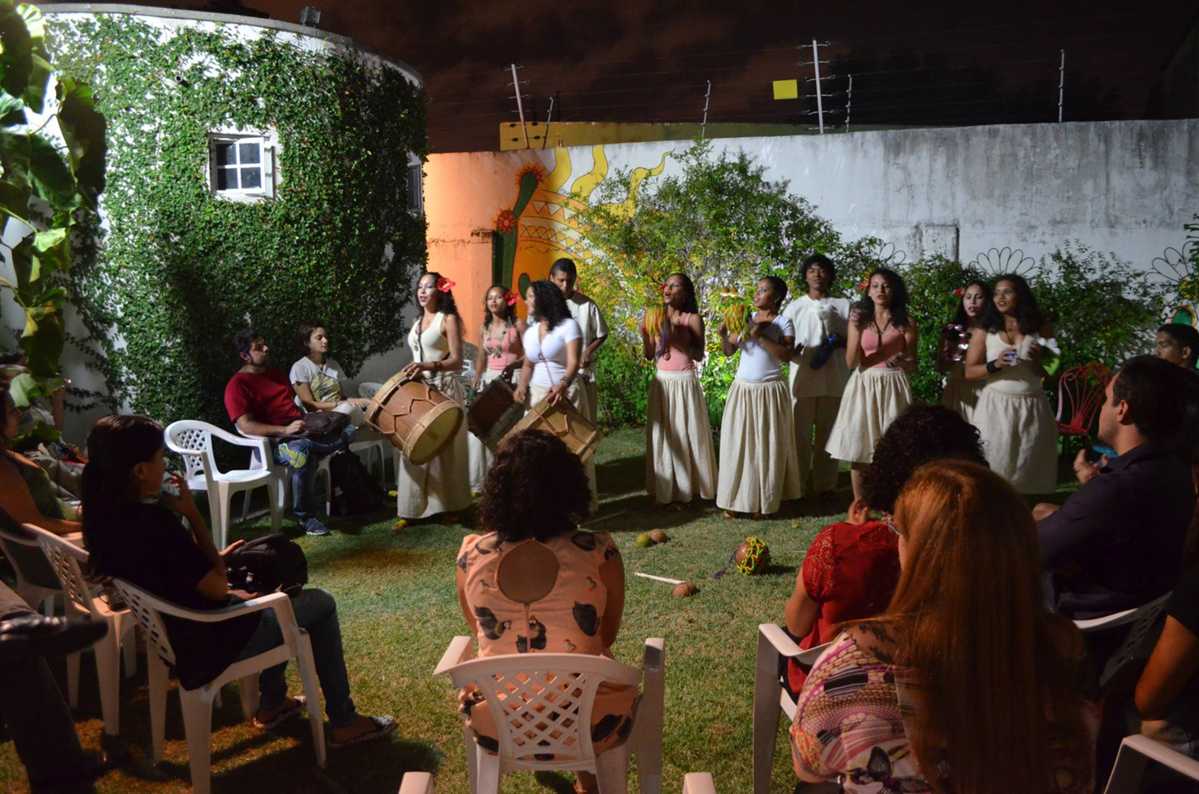 Roda de pessoas sentadas durante a apresentação de grupo em pé com instrumentos