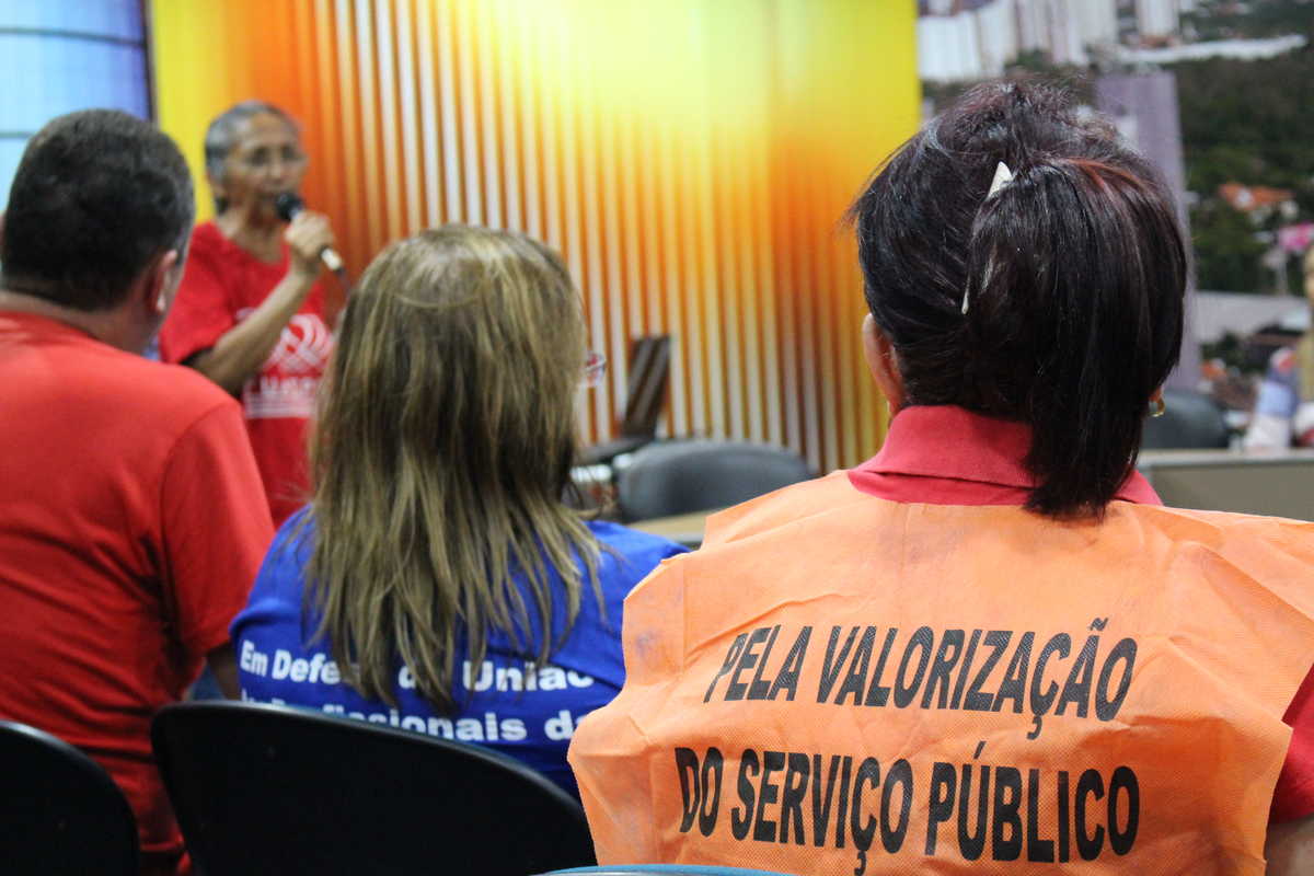 Servidores estaduais reunidos em uma das salas do Complexo das Comissões Técnicas da Assembleia Legislativa do Ceará. No detalhe, uma servidora, sentada, usa colete com a frase 