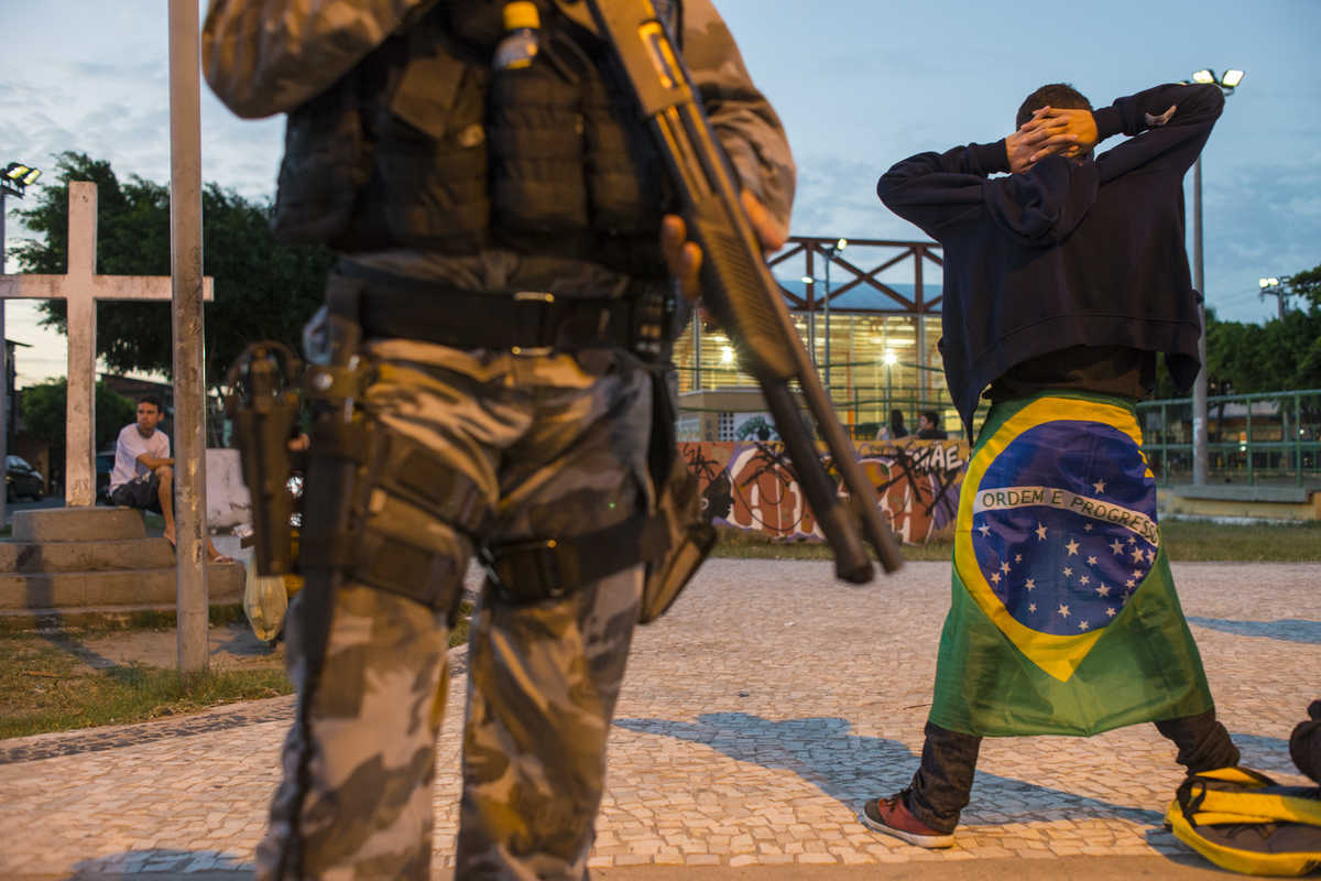 Policial com arma nas mãos e adolescente ao fundo, com as mãos na cabeça e bandeira do Brasil amarrada às costas