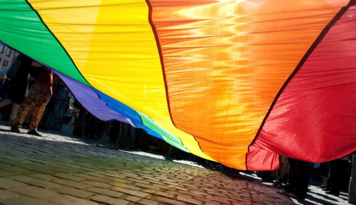 Pés de várias pessoas sob grande bandeira com as cores do arco-íris