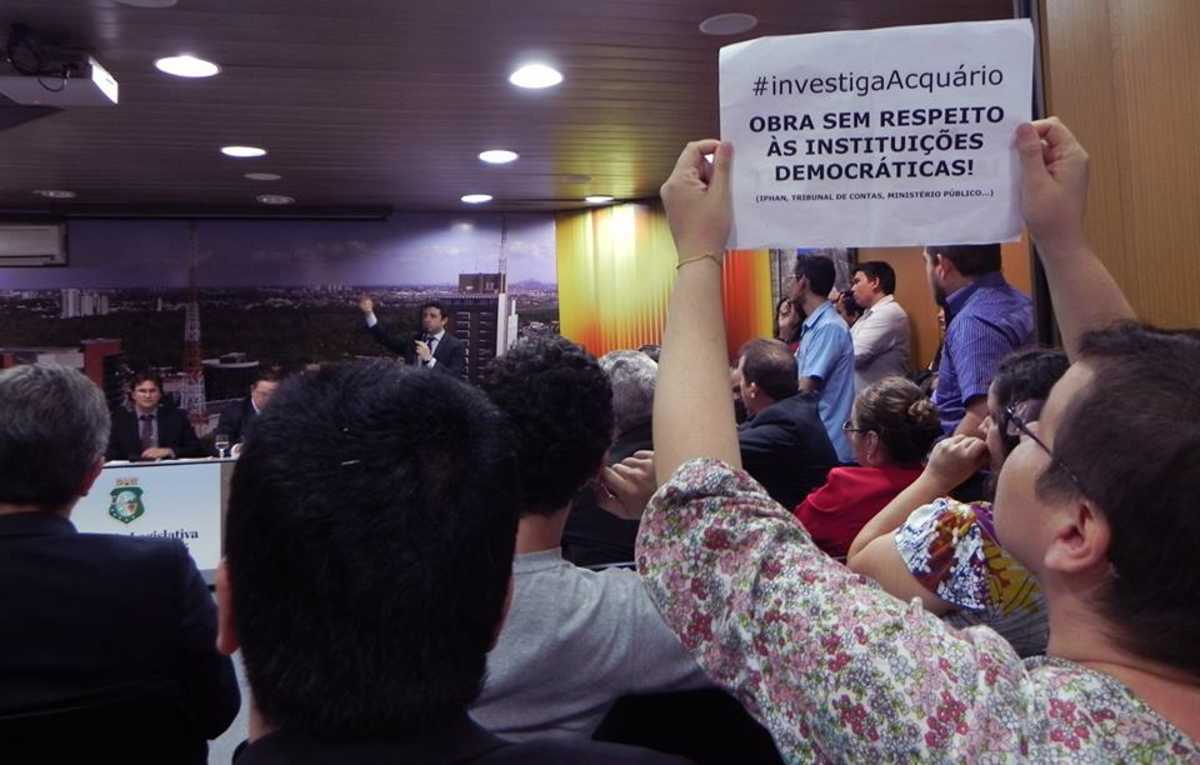 Entre as pessoas que assistem ao debate, uma jovem exibe cartaz com os dizeres 