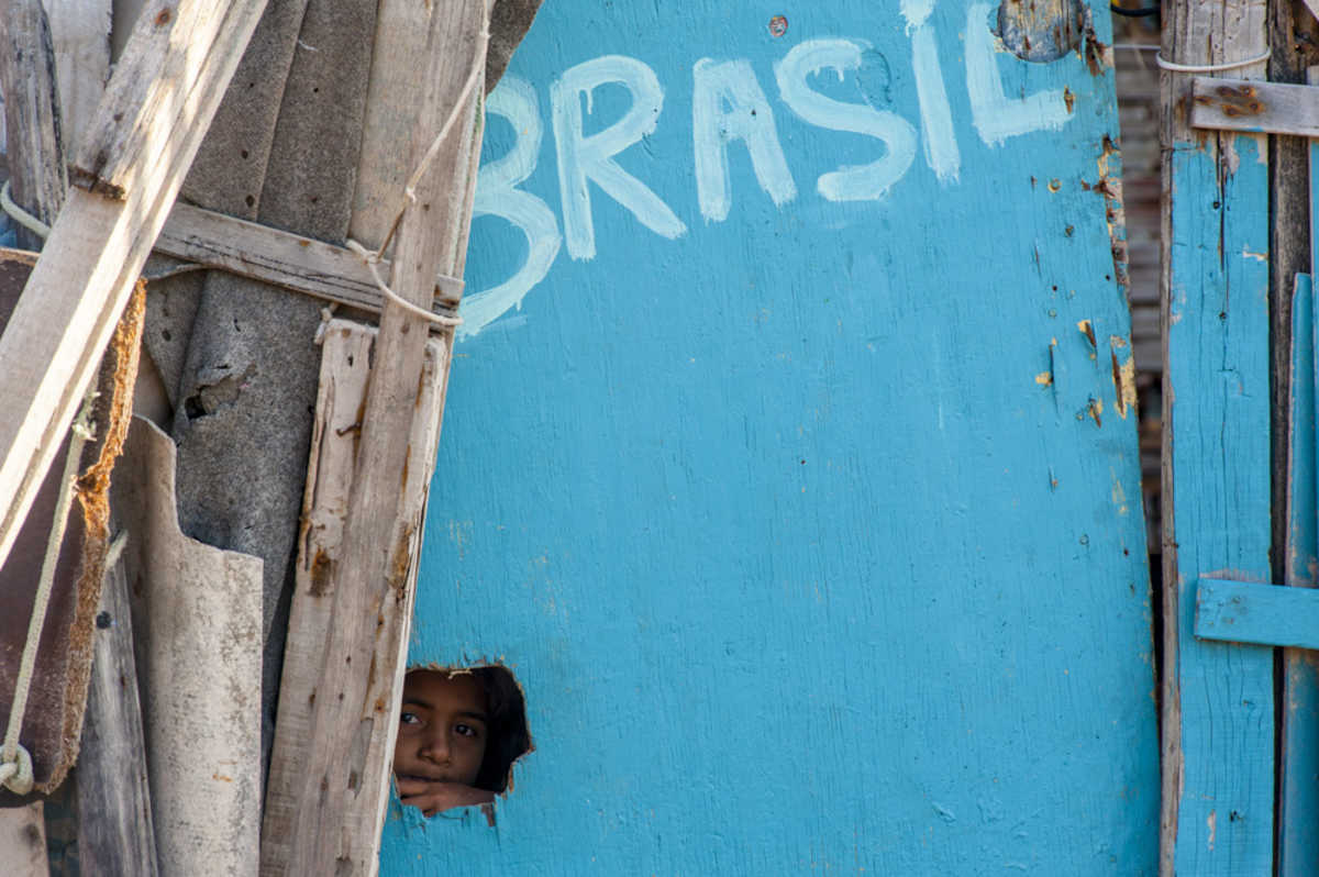 Rosto de criança em buraco de uma moradia precária, contornada por tábuas, onde se lê a palavra Brasil, em cor branca, sobre pintura azul