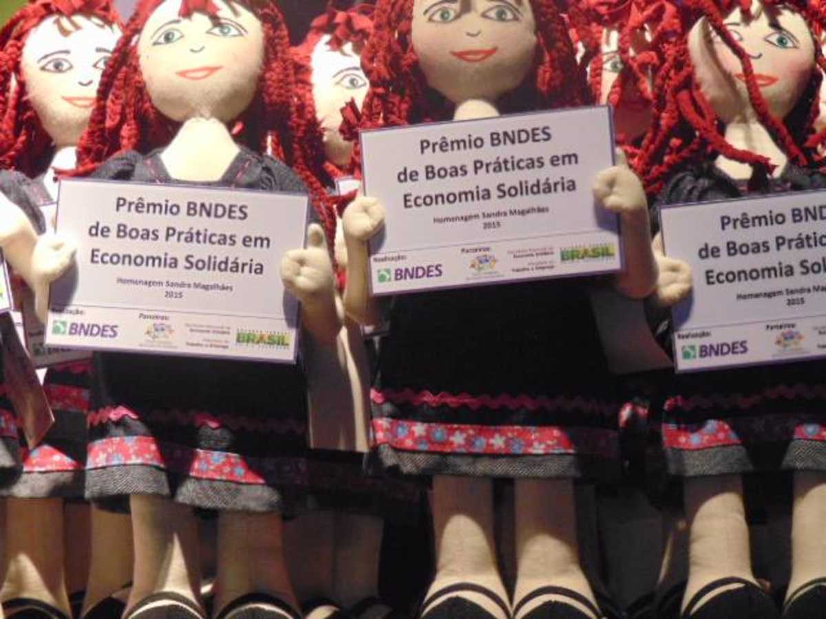 Bonecas de pano seguram cartazes com dizeres sobre prêmio de boas práticas em economia solidária