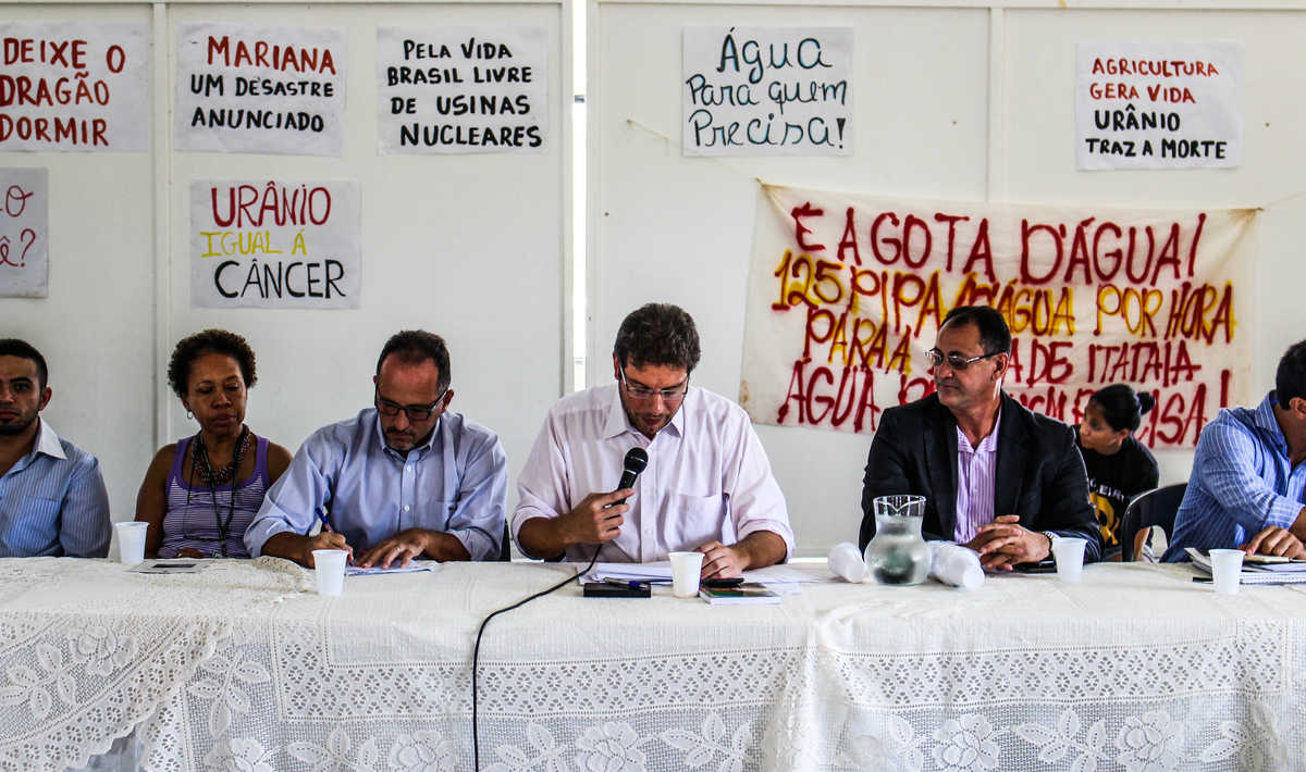 Mesa composta pelo deputado estadual Renato Roseno e outros convidados para a audiência pública, com cartazes ao fundo contra o projeto de mineração