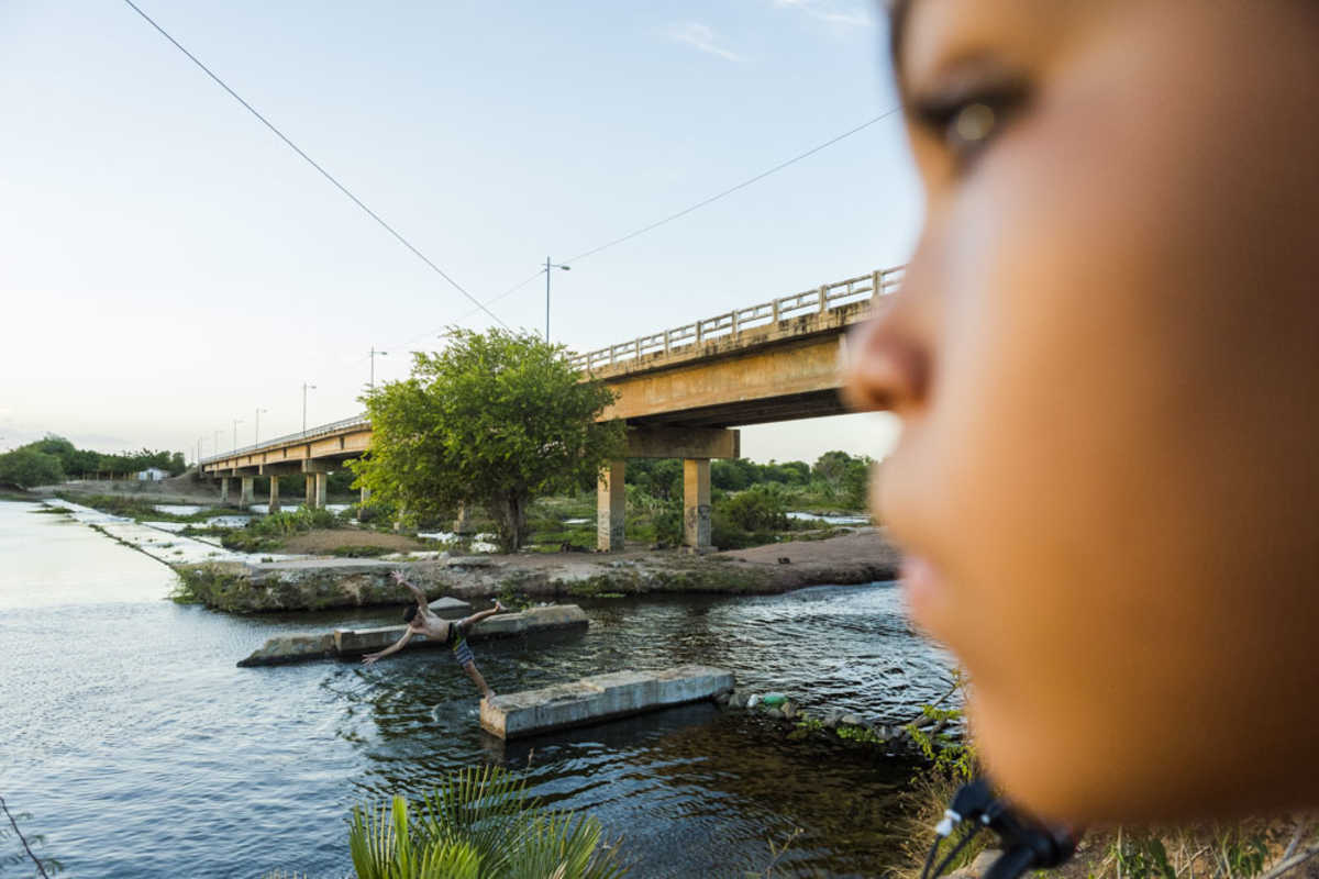 Menino observa outro se jogando nas águas de um rio sob ponte