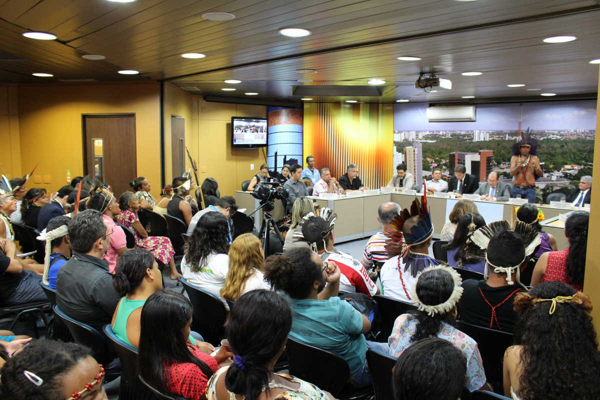 Público da audiência pública na Assembleia Legislativa do Ceará que debateu os direitos dos povos indígenas ouve Weibe Tapeba, coordenador das organizações e povos indígenas, em pé e ao microfone, devidamente caracterizado, na mesa composta para o evento