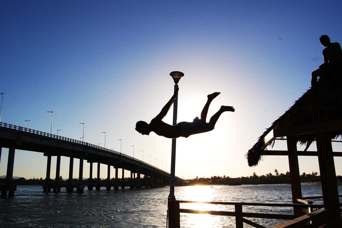 Silhueta de adolescente durante salto em rio, com forte luz ao fundo em contraste com o azul celeste