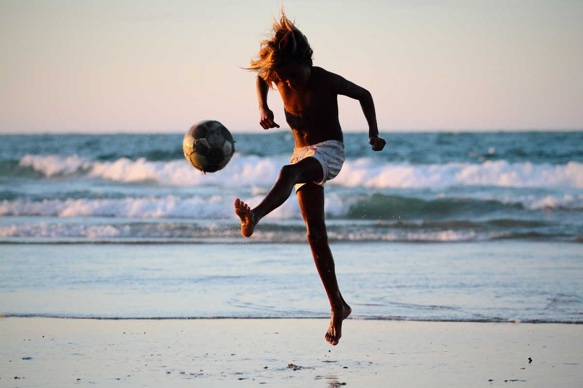 Criança joga bola na areia da praia, com o mar ao fundo